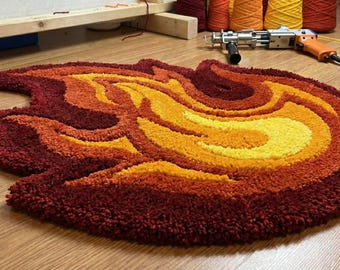 May include: A handmade, flame-shaped rug in shades of red, orange, and yellow. The rug is made with a tufting technique, creating a textured, plush surface. The rug is on a wooden floor.