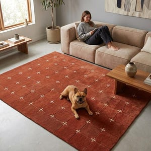 Puede incluir: Una alfombra de color óxido con un patrón de cuadrícula de diseños de puntadas de cruz blancas. Un bulldog francés marrón está acostado en la alfombra. Una mujer está sentada en un sofá beige. Mesas de centro y auxiliares de madera.