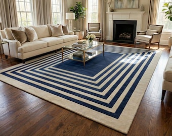 May include: A large area rug with a geometric design. The rug features a navy blue center with cream-colored stripes and border. The rug is placed in a living room with a sofa, chairs, and a coffee table.