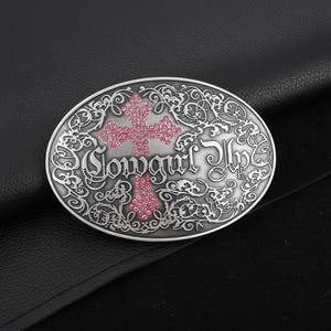 May include: Oval-shaped silver-tone belt buckle with a pink cross design and the words "Cowgirl Up". The buckle has an ornate, swirling border and is set against a black background.