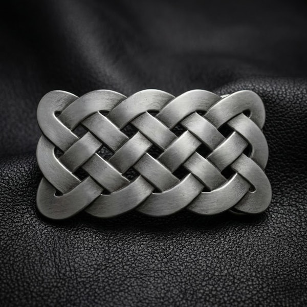 Könnte beinhalten: Silberfarbene Gürtelschnalle mit keltischem Knotenmuster. Die Schnalle hat eine rechteckige Form mit einem ineinander verschlungenen Muster. Sie besteht aus einem metallischen Material. Der Hintergrund ist eine strukturierte schwarze Oberfläche.