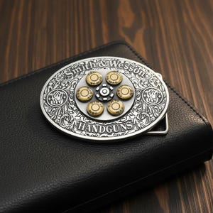 May include: Silver-toned metal belt buckle with a revolver cylinder design. The buckle features the words "Smith & Wesson" and "HANDGUNS" in a decorative font. The center of the buckle has a series of bullet casings. The buckle is resting on a black leather wallet.