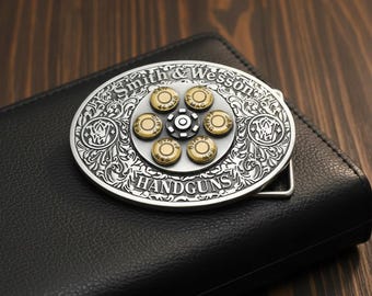 May include: Silver-toned metal belt buckle with a revolver cylinder design. The buckle features the words "Smith & Wesson" and "HANDGUNS" in a decorative font. The center of the buckle has a series of bullet casings. The buckle is resting on a black leather wallet.