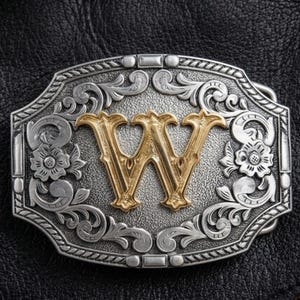 Puede incluir: Hebilla de cinturón de metal plateado con la letra "W" dorada en el centro. La hebilla tiene un diseño floral ornamentado alrededor de los bordes y un fondo texturizado. La hebilla es rectangular y está sobre una superficie de cuero negro.