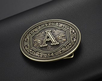 May include: An oval antique brass belt buckle with the letter "A" in an ornate frame. The buckle has an embossed design with a classic western style. The buckle is set against a dark, textured background, with a detailed border.