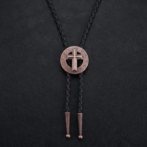 May include: A bronze-colored bolo tie featuring a cross design within a circular frame with an ornate pattern. The tie has a black braided cord with bronze-colored tips and a ball at the end. The background is a textured black surface.