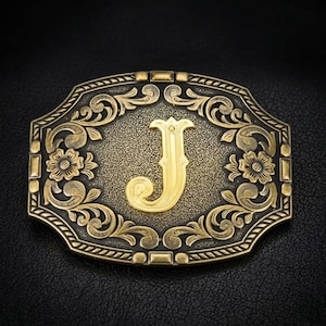 May include: Ornate gold-colored belt buckle with the letter "J" in the center. The buckle features a decorative floral and scroll design around the edges. The background is black.