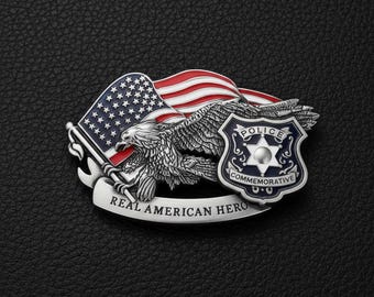 Puede incluir: Una placa de policía plateada con una bandera estadounidense, un águila y una placa de policía. La placa dice "REAL AMERICAN HERO" en una pancarta. La placa de policía es azul y blanca y dice "POLICE COMMEMORATIVE".