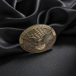 Könnte beinhalten: Ovale Gürtelschnalle aus antikem Messing mit einem Adler mit ausgebreiteten Flügeln und der amerikanischen Flagge. Der Text "THE RIGHT TO BEAR ARMS" ist oben, und "2ND AMENDMENT" darunter. Das Design wird von dekorativen Schnörkeln umrahmt.