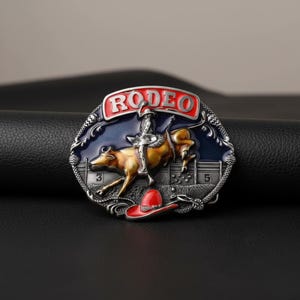 May include: Oval-shaped metal belt buckle with a rodeo theme. The buckle features a cowboy riding a bucking bull, with the word "RODEO" in red. A red cowboy hat is at the bottom. The background is blue and silver.