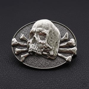 May include: Silver-colored oval belt buckle featuring a detailed skull and crossbones design. The skull has visible teeth and intricate detailing, set against a textured background. The buckle is a classic accessory.