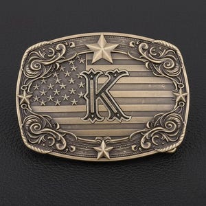 May include: A rectangular, antique brass-colored belt buckle. The buckle features an American flag design with a large letter "K" in the center. The buckle is adorned with stars and decorative scrollwork around the edges.