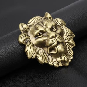 May include: A detailed, antique-style belt buckle featuring a lion's head in a bronze finish. The lion's mane is intricately sculpted, and the buckle is set against a black leather background.