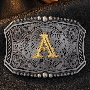 Puede incluir: Hebilla de cinturón de metal plateado y dorado con un diseño floral ornamentado. La hebilla presenta la letra "A" dorada en el centro. La hebilla tiene forma rectangular con bordes redondeados.