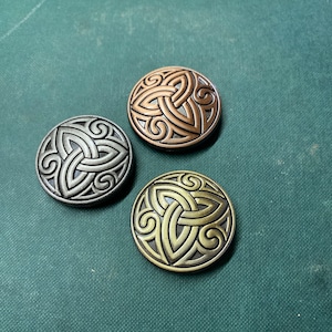 Op de afbeelding: Drie ronde metalen knopen met Keltische knoopontwerpen. Eén knoop is zilverkleurig, één is bronskleurig en één is goudkleurig. De knopen liggen op een donkergroen oppervlak.