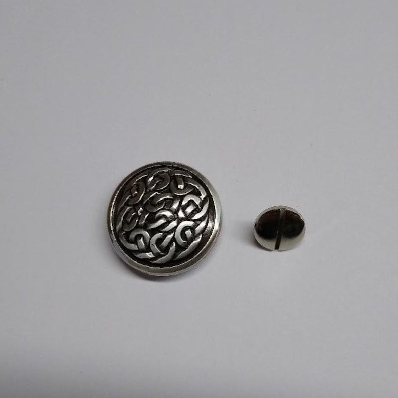 Op de afbeelding: Een zilveren knoop met een Keltisch knoopontwerp. De knoop heeft een zwarte achtergrond en een zilveren rand. De knoop is ongeveer 2,5 cm in diameter.