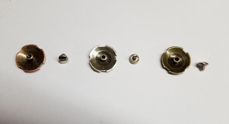 May include: Three sets of metal fasteners, each with a round, decorative top and a screw. The tops are copper, silver, and gold in color.