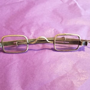 May include: A pair of antique silver-toned reading glasses with a spring hinge and square lenses.