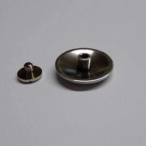 Op de afbeelding: Een zilverkleurige metalen knoop met een verhoogd midden en een schroefdraad aan de onderkant. De knoop is ontworpen om aan een kledingstuk of ander item te worden bevestigd.