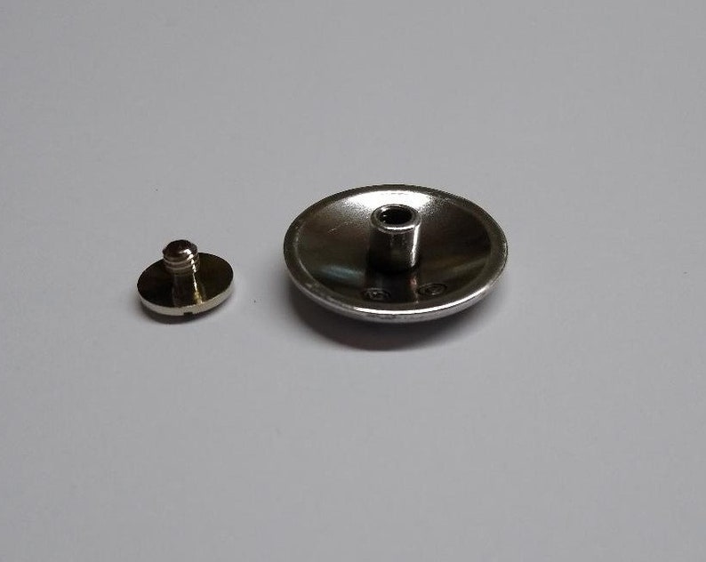 Op de afbeelding: Een zilverkleurige metalen knoop met een verhoogd midden en een schroefdraad aan de onderkant. De knoop is ontworpen om aan een kledingstuk of ander item te worden bevestigd.