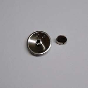Op de afbeelding: Een zilverkleurige metalen knop met een klein gaatje in het midden en een schroef. De knop is rond en heeft een licht verhoogde rand. De schroef is ook zilverkleurig en heeft een platte kop.