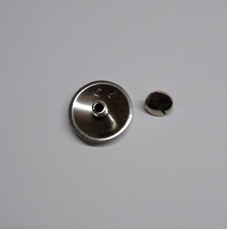 Op de afbeelding: Een zilverkleurige metalen knop met een klein gaatje in het midden en een schroef. De knop is rond en heeft een licht verhoogde rand. De schroef is ook zilverkleurig en heeft een platte kop.