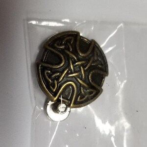 May include: A bronze-colored metal pin with a Celtic knot design. The pin has a circular shape and is attached to a metal backing.