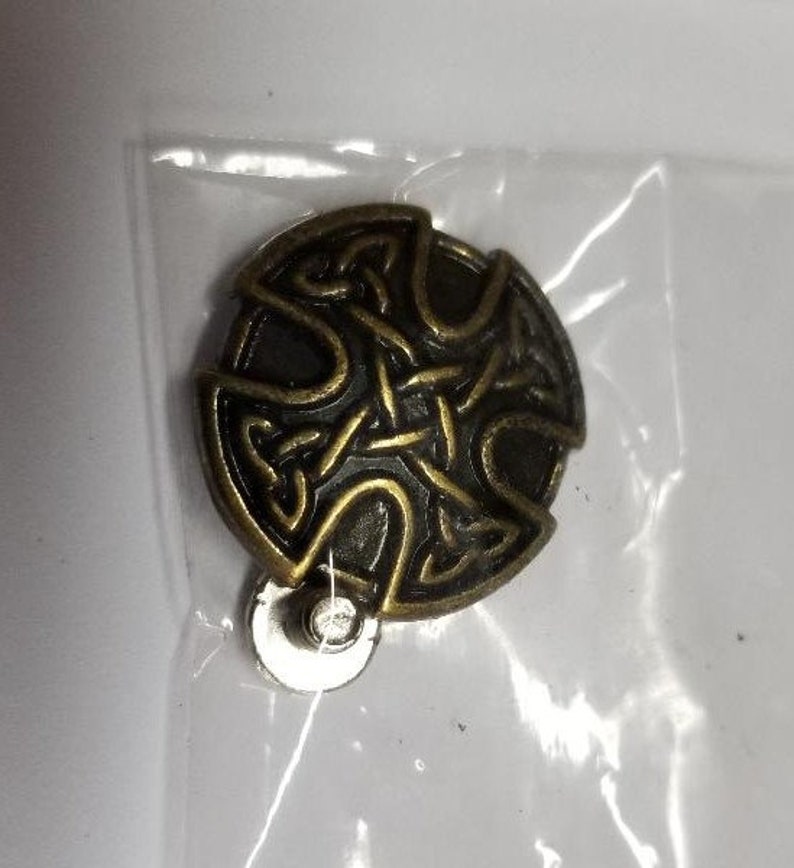 May include: A bronze-colored metal pin with a Celtic knot design. The pin has a circular shape and is attached to a metal backing.