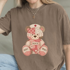 May include: Brown t-shirt with a patchwork teddy bear design. The bear wears a nurse's cap with a red cross and holds a heart that says "NURSE". The bear's paws say "HUGS" and "HEALS".