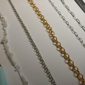 May include: An assortment of jewelry chains in silver and gold tones, alongside a beaded strand. A pink pencil and a teal pen are in the lower left corner. A ruler is in the upper right corner. The chains vary in style, including delicate link and beaded designs.