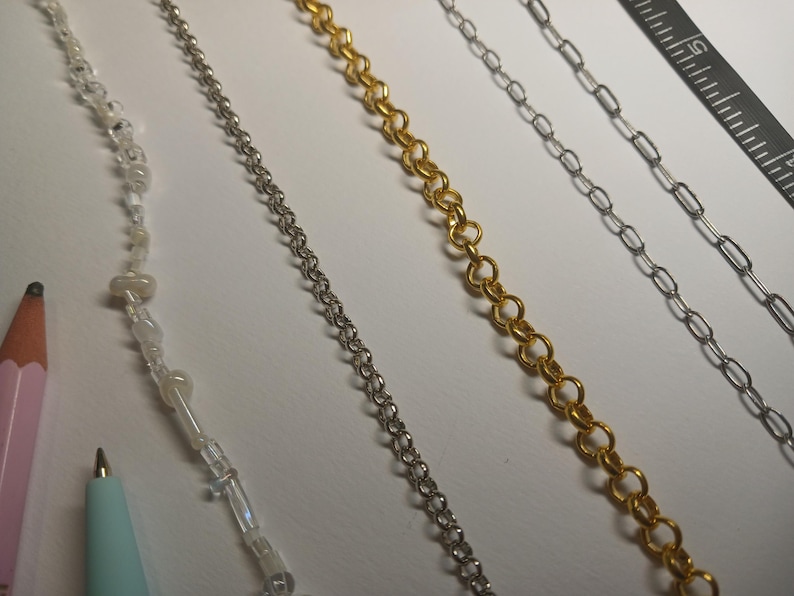 May include: An assortment of jewelry chains in silver and gold tones, alongside a beaded strand. A pink pencil and a teal pen are in the lower left corner. A ruler is in the upper right corner. The chains vary in style, including delicate link and beaded designs.