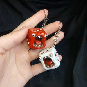 Puede incluir: Dos llaveros con forma de dado, uno rojo y otro blanco, cada uno con una boca muy abierta con dientes y lengua visibles, dos ojos saltones a los lados y pequeños puntos rojos que representan los números del dado con enganche metálico.