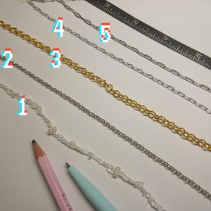 May include: An assortment of jewelry chains in gold and silver tones, including beaded and linked designs. A pink pencil, a blue pen, and a black ruler are also present. The chains are arranged on a white surface.