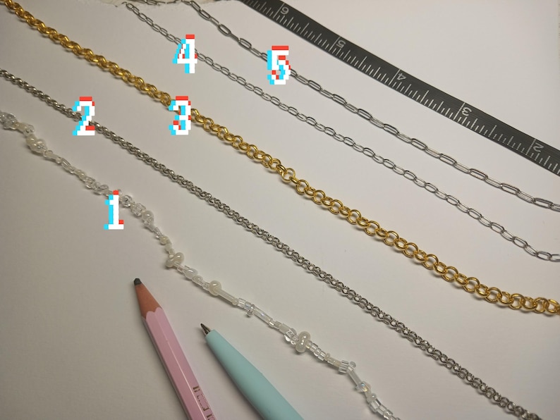 May include: An assortment of jewelry chains in gold and silver tones, including beaded and linked designs. A pink pencil, a blue pen, and a black ruler are also present. The chains are arranged on a white surface.