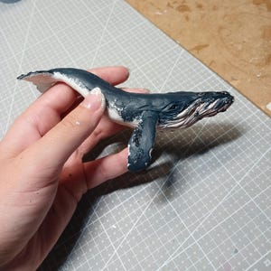 以下が含まれることがあります： 手描きのザトウクジラの置物。クジラは主に濃い青色で、腹部とひれに白いアクセントがあります。置物は灰色のカッティングマットを背景に持っており、海洋哺乳類の彫刻の細部までこだわった職人技が際立っています。