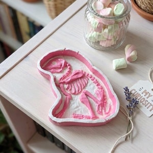 Puede incluir: Un cuenco rosa con forma de esqueleto de flamenco con interior blanco. Se ve un tarro de cristal lleno de malvaviscos rosas y verdes. También hay una ramita de lavanda y una etiqueta que dice "My Curiosity".