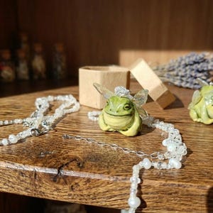 May include: A whimsical scene featuring a green frog pendant with fairy wings, a matching frog figurine, and a delicate beaded necklace. A small open box and a bundle of lavender are also present on a wooden surface.