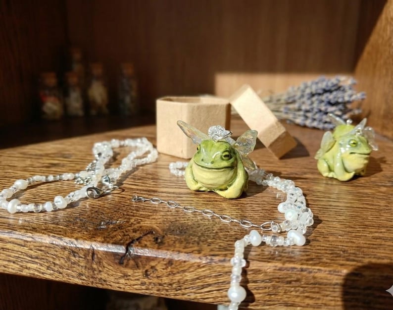 May include: A whimsical scene featuring a green frog pendant with fairy wings, a matching frog figurine, and a delicate beaded necklace. A small open box and a bundle of lavender are also present on a wooden surface.