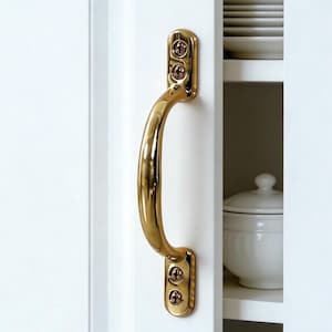 May include: A gold-colored cabinet pull handle mounted on a white cabinet door. The handle has a curved design and is attached with screws. The cabinet door is slightly ajar, revealing white shelves with white dishes and a lidded container.