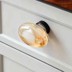 Könnte beinhalten: Ein ovaler Schrankknauf aus bernsteinfarbenem Glas mit schwarzem Sockel ist an einem weißen Schrank befestigt. Der Knauf ist ein dekoratives Hardware-Element, das dem Design des Schranks einen Hauch von Eleganz verleiht. Der Schrank ist Teil einer Küche oder eines Badezimmers.