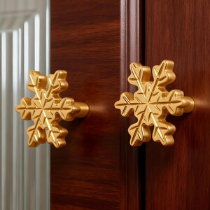Peut inclure: Deux boutons de meuble dorés en forme de flocon de neige fixés sur une porte d'armoire en bois brun foncé. Les boutons présentent des motifs de flocons de neige détaillés. La porte de l'armoire a un panneau de verre avec des lignes verticales.