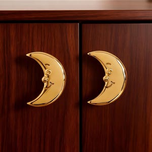 Könnte beinhalten: Zwei goldfarbene Mond-Kabinettgriffe mit lächelnden Gesichtern sind an einem dunklen Holzschrank angebracht. Die Griffe sind symmetrisch und verleihen den Möbeln einen skurrilen Touch.