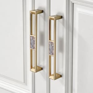 May include: Two gold cabinet pulls with rectangular frames and a central rectangular stone insert. The pulls are mounted on a white cabinet door. The stone insert has a marbled appearance with shades of grey and purple.