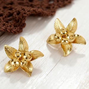 May include: Two gold-colored flower-shaped earrings. Each earring features five detailed petals and a cluster of three spherical accents at the center. The earrings are set against a white wooden surface.