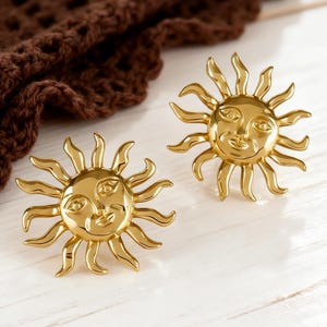 Könnte beinhalten: Zwei goldfarbene, sonnenförmige Ohrringe mit lächelnden Gesichtern und Strahlen. Die Ohrringe liegen auf einer weißen Holzoberfläche, im Hintergrund befindet sich ein braunes Häkeltextil. Die Ohrringe haben eine Vintage- oder Bohème-Ästhetik.