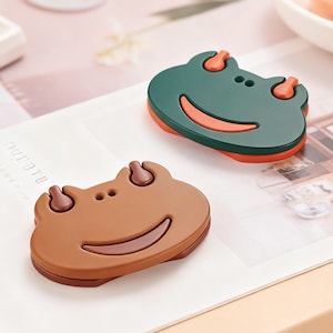 Könnte beinhalten: Zwei froschförmige dekorative Magnete. Einer ist braun mit dunklem braunen Umriss und einem lächelnden Gesicht. Der andere ist grün mit orangefarbenen Akzenten und einem lächelnden Gesicht. Die Magnete liegen auf einer weißen Oberfläche.