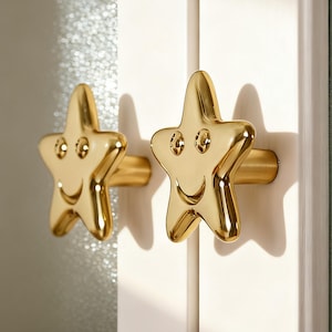 Puede incluir: Dos tiradores dorados en forma de estrella con caras sonrientes. Los tiradores están montados en una puerta de armario blanca. Los tiradores en forma de estrella tienen una superficie brillante y reflectante, añadiendo un toque lúdico.