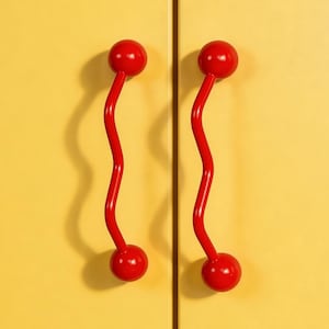 May include: Close-up of a yellow cabinet with two red, wavy handles. Each handle has a red ball at the top and bottom, with a curved, red stem connecting them. The cabinet doors are a solid, matte yellow.