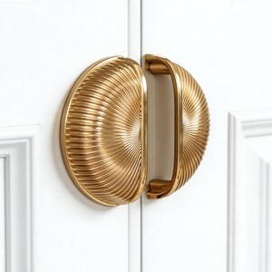 Può includere: Primo piano di due maniglie dorate per armadi. Ogni maniglia ha una forma a semicerchio con un design a coste a forma di ventaglio. Le maniglie sono montate su un'anta bianca, con un semplice design a pannelli incassati.