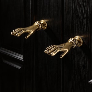 Può includere: Due maniglie dorate a forma di mani, montate su una porta di legno scuro. Le mani sono dettagliate con nocche e unghie visibili. La porta presenta un motivo a venature verticali.